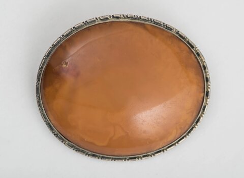 Zdjęcie przedstawia broszę, widok z góry. W centrum owalny bursztyn w miodowym kolorze. Wokół niego obwódka z oksydowanego srebra. 