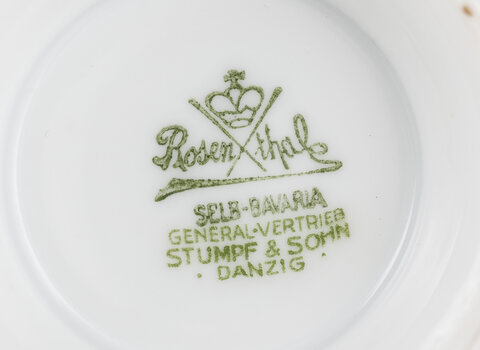 Fragment porcelanowego naczynia przygotowany na cele dobroczynne. Na porcelanie logo producenta Rosenthal (Napis w środku przerwany dwoma liniami w kształcie litery X, powyżej niej korona). Poniżej logo napis "SELB-BAVARIA", niżej "GENERAL-VERTRIEB", niżej "STUMPF & SOHN", niżej "DANZIG".