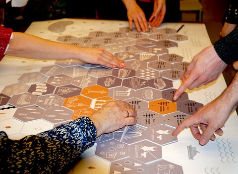 Zdjęcia przedstawia warsztaty Gdańskiej Mozaiki. Dłonie trzymają elementy. Pod nimi plansza z sześciokątów w kształcie obrysu granic administracyjnych współczesnego Gdańska. 