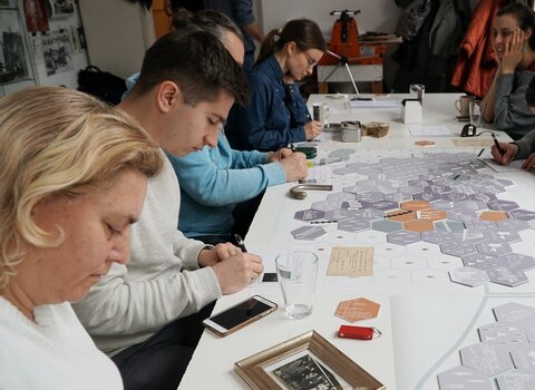 Warsztaty z Gdańskiej Mozaiki odbywają się przy udziale 10 osób. Zdjęcie ukazuje stół a przy nim siedzące osoby z książkami, fotografiami i przedmiotami. 