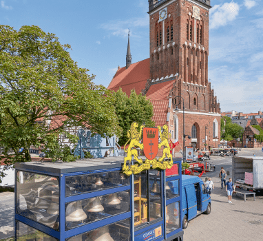 Zdjęcie przedstawia kabinę mobilnego carillonu "Gdańsk". W środku za szybą dzwony. W tle sylwetka kościoła św. Katarzyny.  