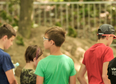 Zdjęcie przedstawia dzieci (po lewej stronie zdjęcia, ukazane od tyłu) patrzące wgłąb wykopu archeologicznego. Po prawej stronie archeolog w kamizelce rozmawiająca z dziećmi. W tle drzewa liściaste. 