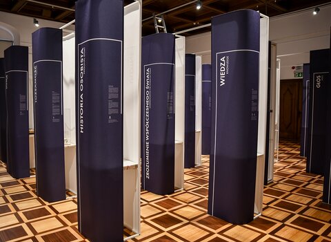 Zdjęcie przedstawia widok na salę wystawową i stojące pylony z napisami. 