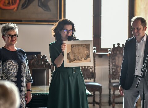 Zdjęcie przedstawia prezydent Gdańska trzymającą stary dokument w oprawie. Po Jej prawej stronie zamyślony Marek Adamkowicz, po lewej uśmiechnięta Halina Pietrzkiewicz. 
