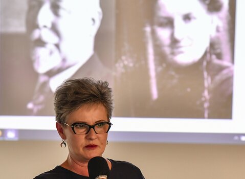 Zdjęcie przedstawia Halinę Pietrzkiewicz trzymającą mikrofon podczas przemowy. W tle archiwalne zdjęcia przedstawiające członków rodziny. 