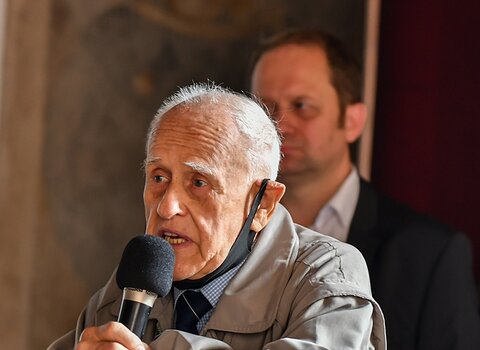 Zdjęcie przedstawia starszego mężczyznę, Kazimierza Bellwona przemawiającego do mikrofonu. Mężczyzna opiera się o krzesło.