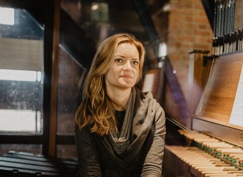 Monika Kaźmierczak siedzi obok klawiatury carillonowej. Blond długie włosy, szczupła sylwetka, uśmiecha się do obiektywu aparatu. 