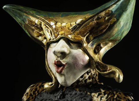 Zdjęcie przedstawia popiersie manekina ubranego w kobiecy wenecki strój karnawałowy. Na głowie maska zakrywająca całą twarz w kolorach zielono-żółto-złotym i zdobiona kawałkami bursztynów. 
Projekt:  Sergio i Massimo Boldrin   Fot. Michał Szczerek 
