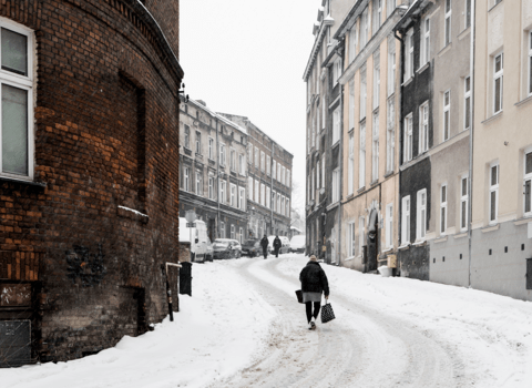 Zdjęcie. Zaśnieżoną ulicą spaceruje kobieta niosąca torby. Po lewej i prawej kamienice na ul. Biskupiej. 