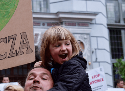 Zdjęcie. Mężczyzna z małą dziewczynką na plecach. Nad nimi transparent z napisem "I Love Puszcza" i sercem. W tle fragment fasady Dworu Artusa. 