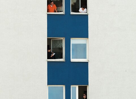 Zdjęcie. Biała ściana budynku. W prawej części niebieska kolumna, a wewnątrz niej pięć rzędów okien, w każdym po dwa okna. W oknach ludzie w różnych pozach. 