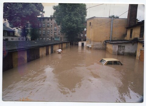 Zdjęcie zalanego wodą dziedzińca. Po lewej, wzdłuż garaży, w wodzie brodzi człowiek. Po prawej z mętnej wody wystaje dach fiata 126. W tle budynki.  