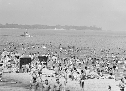 Fragment czarno-białej fotografii. Na zdjęciu plaża na gdańskich Stogach. Na brzegu i w wodzie liczni ludzie. W tle fragment brzegowy w kierunku Jelitkowa. Na morzu niewielki statek wycieczkowy. 