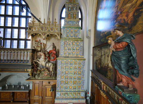 Zdjęcie przedstawia wnętrz Wielkiej Hali Dworu Artusa. W centrum narożnik Hali z Wielkim Piecem oraz rzeźbą św. Jerzego zabijającym smoka pod gotyckim sklepieniem. 10-metrowy piec sięga sklepień.