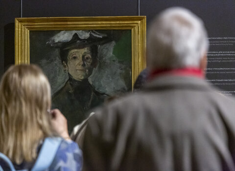 Wernisaż wystawy Olgi Boznańskiej. Zdjęcie przedstawia dwie osoby, mężczyznę i kobietę (po prawej) stojących plecami. Za nimi w tle fragment autoportretu artystki. 