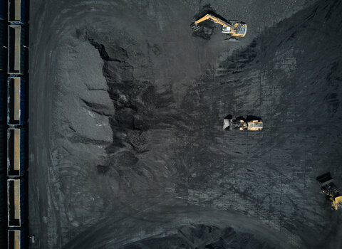 Zdjęcie z drona. Widok na nabrzeże węglowe. Wszystko jest czarne. Wyróżniają się tylko trzy żółte koparki, oraz kontenery stojące w rzędzie na nabrzeżu, po lewej stronie zdjęcia. Koparki zagarniają węgiel na wielką hałdę.
