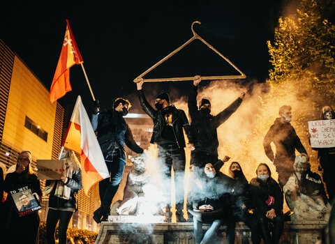 Zdjęcie wieczorne z protestu. 10 osób w maseczkach ochronnych stoi, trzymają transparenty z hasłami przeciw partii rządzącej i zaostrzeniu prawa aborcyjnego. Dwaj mężczyźni pośrodku trzymają w górze wielki wieszak. Za nimi unosi się smuga dymu.
