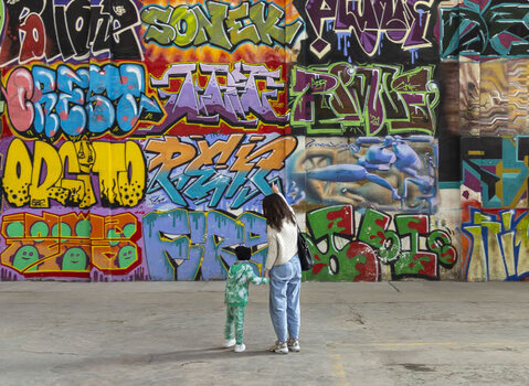 Zdjęcie. Kobieta z dzieckiem stoją przed wysoka na kilkanaście metrów ścianą. Cała zamalowana jest kolorowymi graffiti. Ścianę podzielono na 48 pól, każde wypełnia inne graffiti.