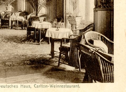 Zdjęcie czarno-białe. Wnętrze budynku. Liczne stoły nakryte obrusami i krzesła. Napis w języku niemieckim informuje, że to wnętrze restauracji hotelu Deutsches Haus.  