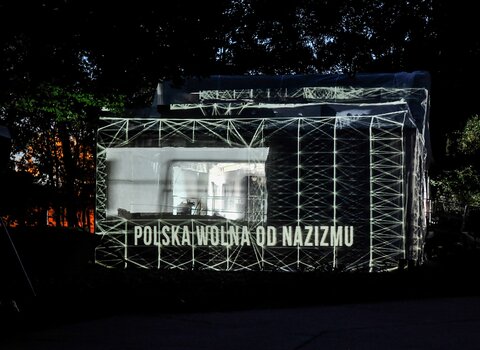 Zdjęcie przestawia bryłę Nowych Koszar na Westerplatte otoczonej przez drzewa. Budynek jest przykryty materiałem. Na nim są wyświetlane geometryczne wzory. Poniżej napis "Polska wolna od nazizmu". 