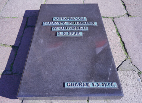 Zdjęcie przedstawia tablicę wmurowaną w posadzkę placu Obrońców Poczty Polskiej. Na kamieniu napis Obrońcom Poczty Polskiej w Gdańsku 1.9.1939 oraz Gdańsk 1.9.46