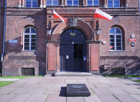 Zdjęcie przedstawia fasadę i drzwi wejściowe do budynku. Nad wejściem po lewej i prawej stronie zwisają polskie flagi. Bo bokach okna. 