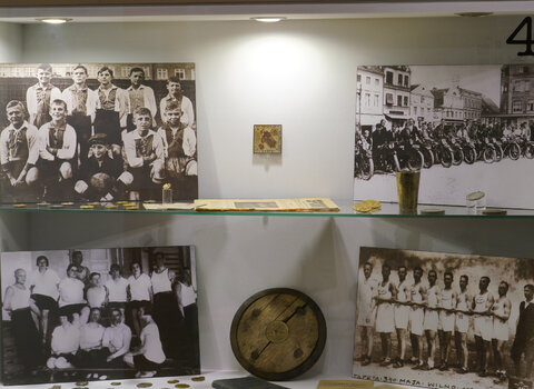 Zdjęcie przedstawia gablotę na salach wystawowych. W środku dysk, medale oraz cztery zdjęcia ukazujące sportowców - dzieci, dorosłych na rowerach i kobiet. 