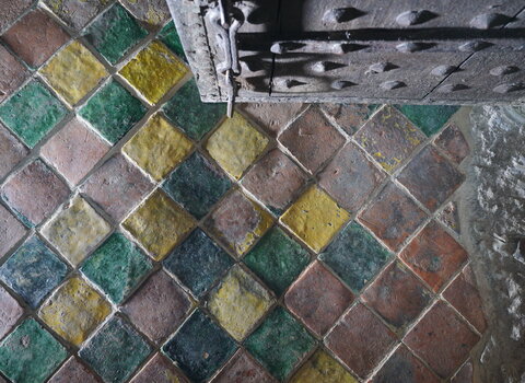 Zdjęcie przedstawia podłogę w pomieszczeniu Wielki Krzysztof. Płytki mają kształt kwadratów. Mają złote, turkusowe oraz ceglane kolory. W nieregularnej mozaice dominują te ostatnie. W górnej części zdjęcia drzwi.  