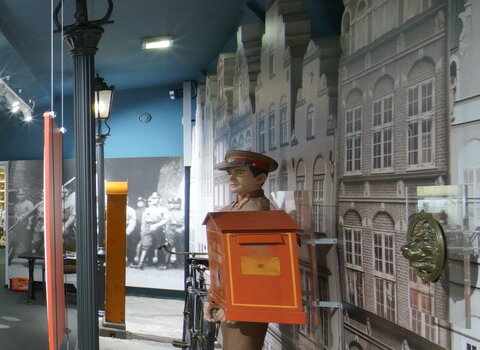 Zdjęcie przedstawia salę wystawową. Po lewej pomalowana na biało-czerwono budka wartownicza z okresu Wolnego Miasta Gdańska. W tle fasady kamieniczek oraz słupy ogłoszeniowe z plakatami z lat 20-30 XX w. i szklane galerie sklepów oraz odtworzonych pomieszczeń mieszkalnych.