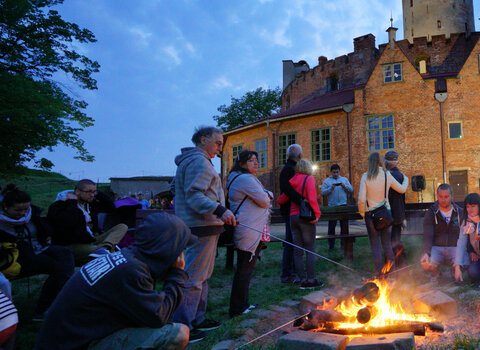 Zdjęcie przedstawia na pierwszym planie grupę osób przy ognisku na dziedzińcu Twierdzy, w tle wieża i otaczające ją kamieniczki. 