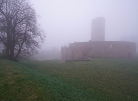 Zdjęcie przedstawia widok na Twierdzę Wisłoujście z dali. Po prawej sylwetka wieży i otaczających kamieniczek widoczna we mgle, po lewej stronie drzewo. 