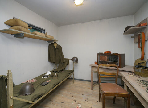 Zdjęcie przedstawia salę wystawową we wnętrzu Wartowni odtwarzającej pomieszczenie, w którym przebywali żołnierze. Po lewej stronie widoczne łóżko polowe, po lewej dwa drewniane stoły, na których stoją radio i łącznica. Na meblach rozmieszczone elementy umundurowania i uzbrojenia polskich żołnierzy. 