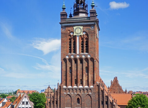 Widok z oddali na kościół św. Katarzyny, w którym mieści się Muzeum Nauki Gdańskiej. Widok fasady budynku. Na wieży kościoła umieszczony okrągły zegar, wieża zwieńczona balustradą i wieżyczkami. 