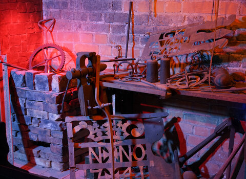 Zdjęcie przedstawia aranżację warsztatu kowalskiego. Na półkach leżą liczne narzędzia, np. obcęgi, młoty i szczypce.