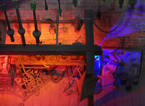 Zdjęcie przedstawia aranżację warsztatu kowalskiego widoczną z góry. Na półkach leżą liczne narzędzia, np. obcęgi, młoty i szczypce.