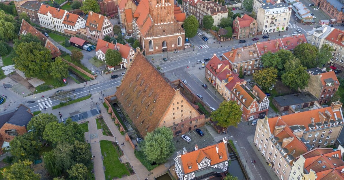Zdjęcie przedstawia zabudowę Gdańska, Wielki Młyn z lotu ptaka. Z dużej wysokości widać trójkątny dach Wielkiego Młyna, Kościół św. Katarzyny, inne ceglane budynki i tereny zielone. 
