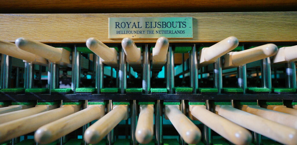 Klawiatura carillonu. Dwa rzędy drewniane pałek pełniących funkcję klawiszy. Nad nimi tabliczka znamionowa holenderskiej firmy Royal Eisbouts - producenta instrumentu. 
