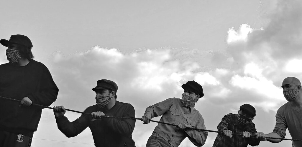 Zdjęcie przedstawia grupę mężczyzn trzymających naprężoną linę. Odtwarzają powojenne zdjęcie robotników pracujących przy odgruzowywaniu Gdańska.  