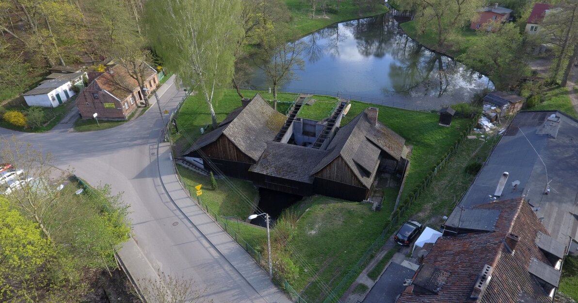 Zdjęcie wykonane z drona przedstawia budynek Kuźni Wodnej wraz z otoczeniem. W centralnym miejscu jest budynek kuźni, po lewej i prawej sąsiadujące budynki,  w tle zbiornik wodny na Potoku Oliwskim oraz Trójmiejski Park Krajobrazowy. 
