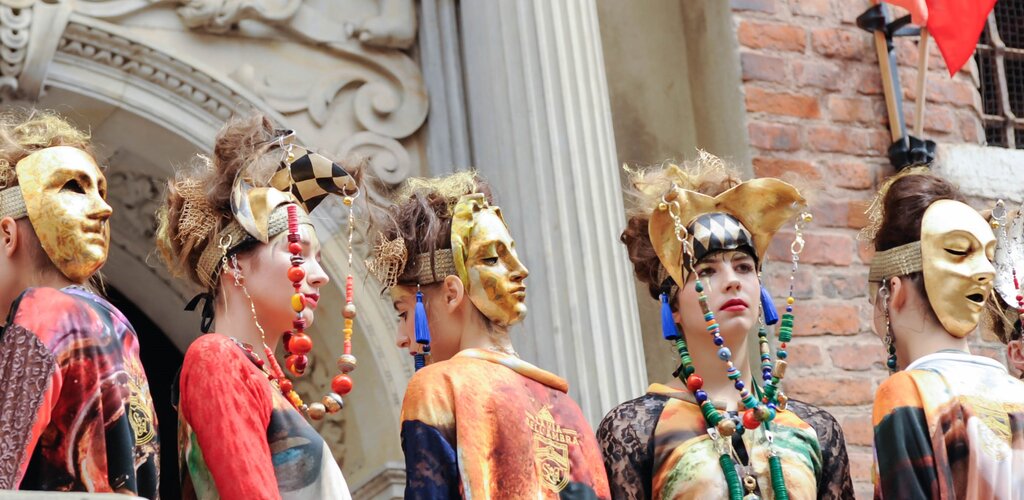 Na zdjęciu zrobionym z dołu znajduje się pięć kobiet ubranych w kolorowe stroje. Do ozdób na ich głowach są przymocowane długie barwne korale. Trzy z nich są odwrócone plecami do widza, na tyłach głów mają założone złote maski przedstawiające twarze. 
