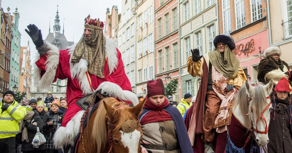 Mężczyźni przebrani za trzech króli jadą na koniach pod ul. Długiej i pozdrawiają przechodniów, za nimi widać tłum osób.  