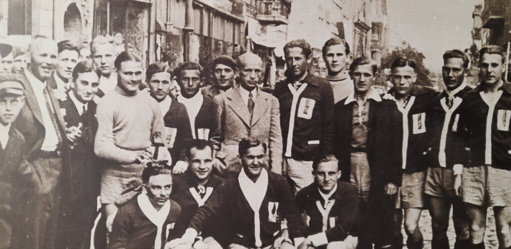 Czarno-białe zdjęcie z ok. 1932 roku. Członkowie sekcji piłkarskiej wraz z władzami klubu i sympatykami pozują do zdjęcia wśród toruńskich kamienic. 