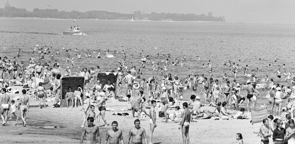 Fragment czarno-białej fotografii. Na zdjęciu plaża na gdańskich Stogach. Na brzegu i w wodzie liczni ludzie. W tle fragment brzegowy w kierunku Jelitkowa. Na morzu niewielki statek wycieczkowy. 
