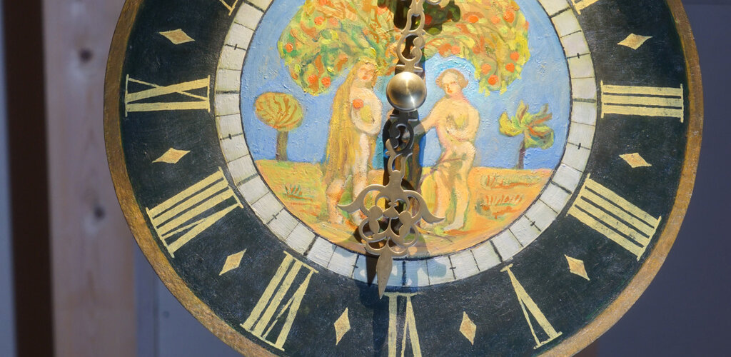 Zdjęcie przedstawia tarczę zegara żuławskiego. Na okrągłej tarczy namalowano w kolorze scenę z biblijnego raju – Ewę i Adama stojących pod drzewem. Rysunek otacza czarna obwódka, na której umieszczonych jest dwanaście cyfr rzymskich wskazujących godziny. 