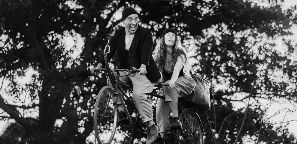 Zdjęcie czarno białe. Spektakl uliczny. Mężczyzna i kobieta siedzą na rowerze dla dwóch osób, tandemie. Rower unosi się w powietrzu, zawieszony na linkach.