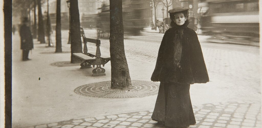 Olga Boznańska na ulicy miasta. W tle ulica, drzewa i ławki. 