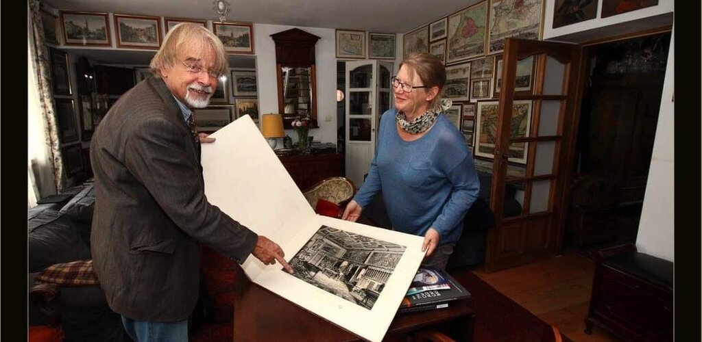 Zdjęcie. Profesor Heinsohn z żoną Joanną, trzymają razem otwarty album, z czarno-białym zdjęciem zabytkowego gdańskiego pomieszczenia. Uśmiechają się. Stoją w pokoju, na ścianach wiszą mapy i zabytkowe grafiki z gdańskimi widokami.